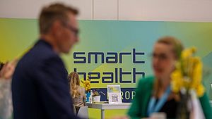 Logo der Smart Health Europe auf einer Wand, im Vordergrund Menschen im Gespräch, unscharf und nicht erkennbar