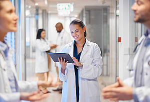 Ärztin auf Krankenhausflur mit Tablet in der Hand, um sie herum andere Mitarbeiter:innen, die sich miteinander unterhalten