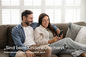 Paar sitzt auf Sofa mit Handy in der Hand der Frau mit Schriftzug "Sind beim Kinderwunscharzt."