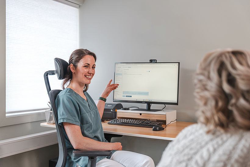 Eine lächelnde Ärztin sitzt mit einer Patientin vor Bildschirm und zeigt mit Finger auf den Screen.